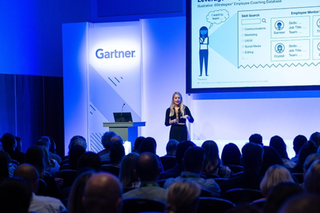 A speaker presents on stage at a Gartner conference while attendees watch from a seated audience.